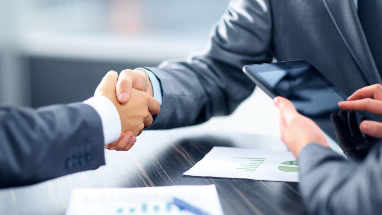 Deux professionnels en costume se serrant la main, avec une tablette et des documents financiers sur la table, illustrant l'importance du reporting financier pour une gestion efficace.